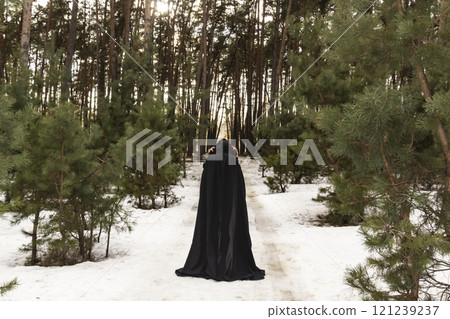 Blonde woman in a black cloak with a hood in winter in a pine forest. Female in cloak with hood from the view from the back side. Blonde woman in a black cloak with a hood in winter in a pine forest. Female in cloak with hood from the view from the back side. 121239237