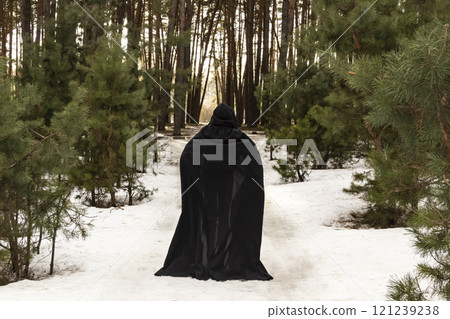 Blonde woman in a black cloak with a hood in winter in a pine forest. Female in cloak with hood from the view from the back side. Blonde woman in a black cloak with a hood in winter in a pine forest. Female in cloak with hood from the view from the back side. 121239238