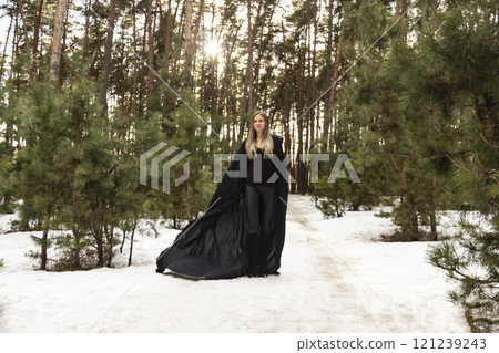 Blonde woman in a black cloak with a hood in winter in a pine forest 121239243