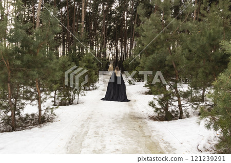 Blonde woman in a black cloak with a hood in winter in a pine forest. Female in cloak from the view from the back side 121239291