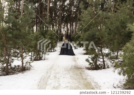 Blonde woman in a black cloak with a hood in winter in a pine forest. Female in cloak from the view from the back side Blonde woman in a black cloak with a hood in winter in a pine forest. Female in cloak from the view from the back side 121239292