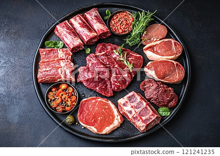 Variety of raw red meat cuts with herbs and spices on black plate 121241235