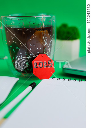 A mug of tea with a red label where it says 2025 stands in the center of the image on a green background, behind it is a green moss flower in a white pot, you can see a banknote with a white pencil, a A mug of tea with a red label where it says 2025 stands in the center of the image on a green background, behind it is a green moss flower in a white pot, you can see a banknote with a white pencil, a 121243280