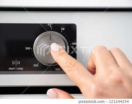 Woman adjusting modern white oven in kitchen.  Female hand pushing and turning knobs on oven control panel 121244254