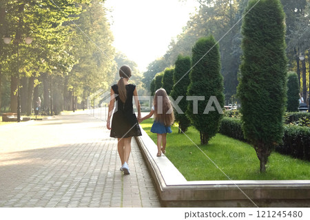 Mother and her small daughter with long hair walking together holding hands in summer park. 121245480