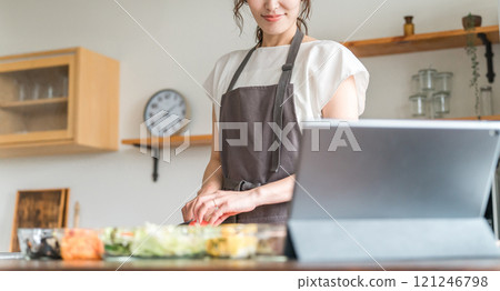 A woman in an apron cooking while looking at a recipe using an app on a tablet 121246798