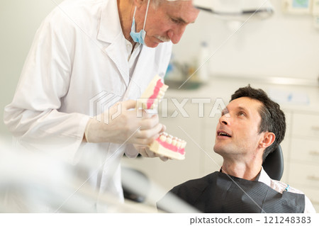 Senior man dentist communicates with male patient, shows model of jaws Senior man dentist communicates with male patient, shows model of jaws 121248383
