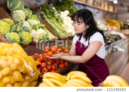 Young saleswoman placing tomatos on food stall in grocery store 121248583