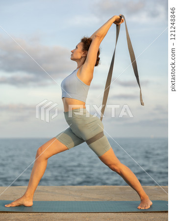 Yoga practice on embankment, female athlete fulfil carry out warrior pose with belt 121248846