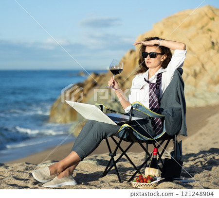 Businesswoman working on laptop and drinking red wine on seashore. Remote work concept Businesswoman working on laptop and drinking red wine on seashore. Remote work concept 121248904
