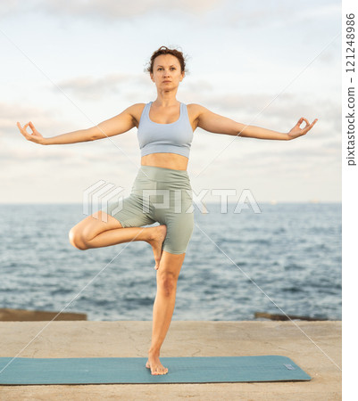 Athletic female yoga practitioner performing Garudasana on beach Athletic female yoga practitioner performing Garudasana on beach 121248986