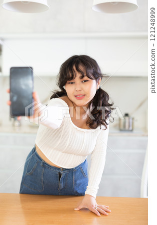 Smiling young girl showing modern smart phone with blank screen, in the living room or in kitchen Smiling young girl showing modern smart phone with blank screen, in the living room or in kitchen 121248999
