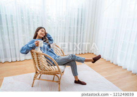 Young Asian woman happy and smiling, sitting on seat and holding mug in living room at home Young Asian woman happy and smiling, sitting on seat and holding mug in living room at home 121249039