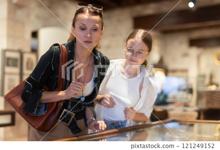 Young woman with teen daughter visitors stands near horizontal glass display case 121249152