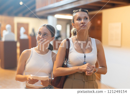 Young woman with teen daughter walk around museum, view sculpture in museum hall 121249154