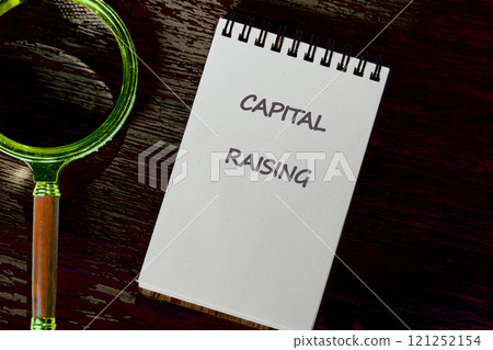 Notepad displaying capital raising notes beside a magnifying glass on a wooden surface 121252154