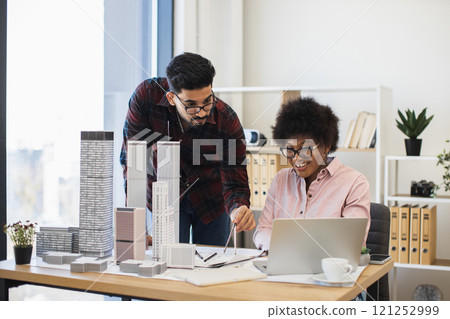 Indian man and African woman architects focused on innovative city project at desk. Collaborative teamwork in office setting with architectural models and laptop. 121252999