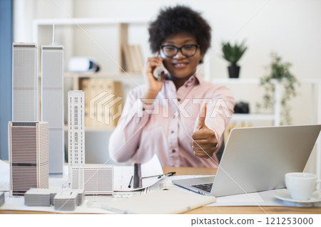 Confident African businesswoman architect having phone conversation about city project. Smiling and giving thumbs up while working with laptop and architectural models in office. 121253000