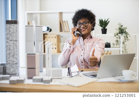Confident African businesswoman architect having phone conversation about city project. Smiling and giving thumbs up while working with laptop and architectural models in office. 121253001