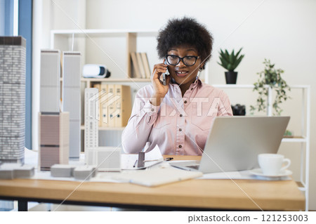 African American woman architect in modern office discussing city project on smartphone, surrounded by building models. Professional setting conveys creativity, architecture, communication. African American woman architect in modern office discussing city project on smartphone, surrounded by building models. Professional setting conveys creativity, architecture, communication. 121253003