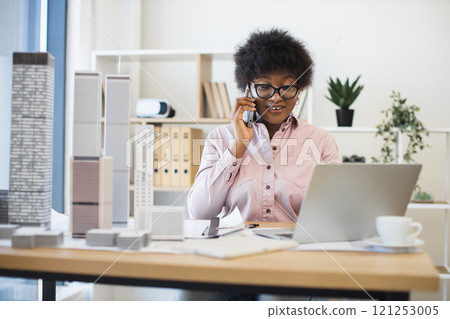 African American woman architect in modern office discussing city project on smartphone, surrounded by building models. Professional setting conveys creativity, architecture, communication. 121253005