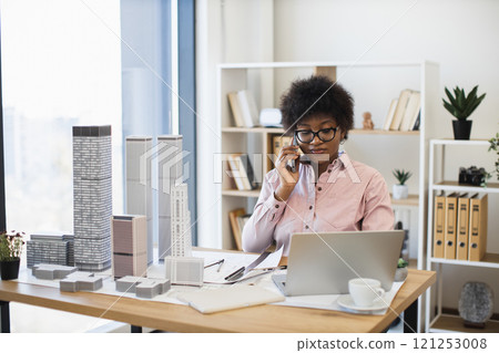 Confident African woman architect discussing city project on smartphone. Office setting with skyscraper models, laptop, and paperwork. Young professional working on modern urban design. 121253008
