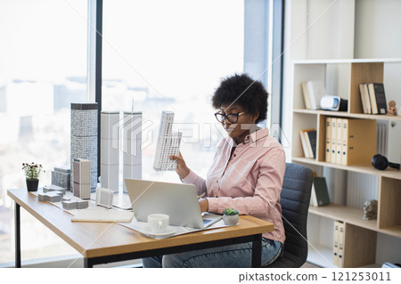 African woman architect in pink shirt designs modern city model at office. Creative workspace features architectural models, laptop, and stationery. African woman architect in pink shirt designs modern city model at office. Creative workspace features architectural models, laptop, and stationery. 121253011