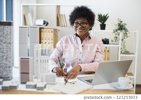 African woman architect in office drawing modern city project. Young professional focused on plans surrounded by architectural models and laptop. African woman architect in office drawing modern city project. Young professional focused on plans surrounded by architectural models and laptop. 121253015