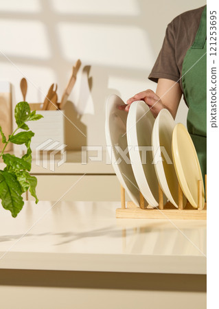 A template picture about kitchen concept, frontal view of a housewife is touching a plate placed on a wooden holder, on a beige surface, next to a branch, and behind have some kitchen supplies. 121253695