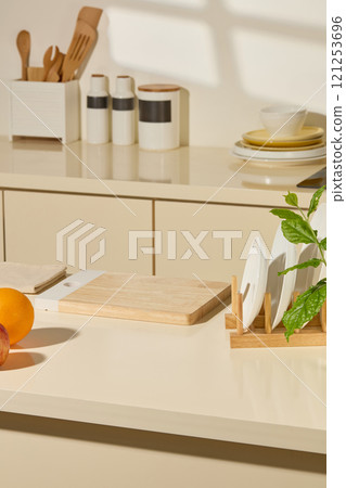 A kitchen-themed photograph features a wooden cutting board in the center of a table, plates on a wooden holder, fruits, spice jars, and wooden kitchen utensils in a white box. Frontal view. 121253696
