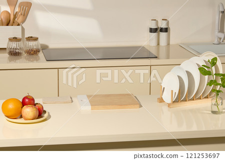 A picture with a kitchen theme shows a wooden cutting board in the middle of a table, plates on a wooden holder, a plate of fruits, spice jars, and a white box filled with wooden kitchen utensils. A picture with a kitchen theme shows a wooden cutting board in the middle of a table, plates on a wooden holder, a plate of fruits, spice jars, and a white box filled with wooden kitchen utensils. 121253697