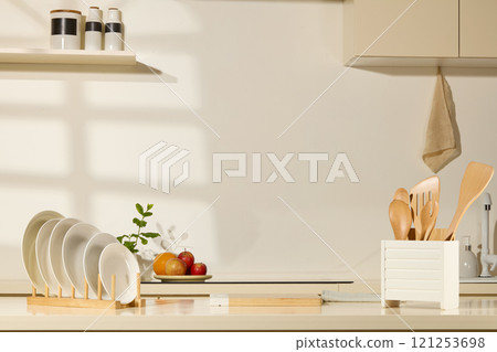 In the bright kitchen background, a wooden cutting board sits in the center of a table, next to a wooden holder, plates, a white box, wooden cooking equipment, fruit plates, and spice jars. 121253698