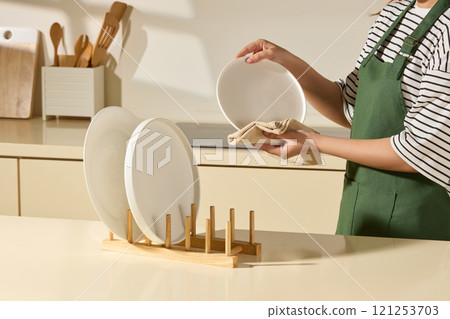 A close-up view of a corner of the kitchen, a housewife wiping a plate, while some plates placed on a wooden holder, a white receptacle containing wooden supplies, next to a cutting board is behind. 121253703