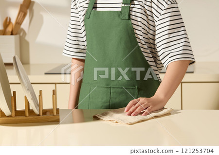 Background in a cozy kitchen, a frontal angle of a housewife is wiping a kitchen counter; next to a wooden holder of plates, behind her is a white receptacle containing wooden kitchen utensils. 121253704
