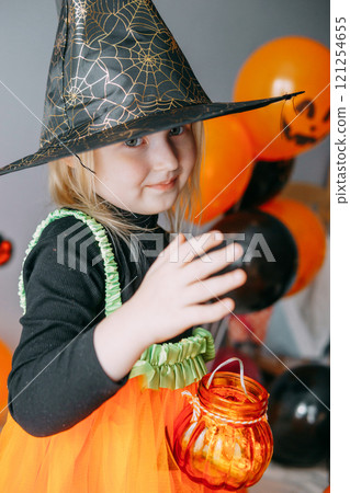 Children's Halloween - a girl in a witch hat and a carnival costume with airy orange and black balloons at home. Ready to celebrate Halloween 121254655