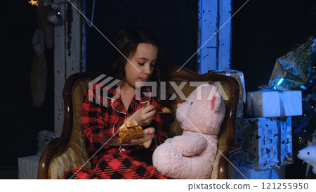 Cute little girl in night pajamas eats a festive cake while sitting in a chair near a Christmas tree. The girl treats her favorite toy, a pink teddy bear, with cake. The concept of a family Cute little girl in night pajamas eats a festive cake while sitting in a chair near a Christmas tree. The girl treats her favorite toy, a pink teddy bear, with cake. The concept of a family 121255950