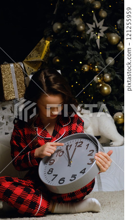 Little girl in night pajamas holding a watch in her hands waiting for Santa Claus and magic. Christmas night, festive atmosphere. Little girl in night pajamas holding a watch in her hands waiting for Santa Claus and magic. Christmas night, festive atmosphere. 121255959