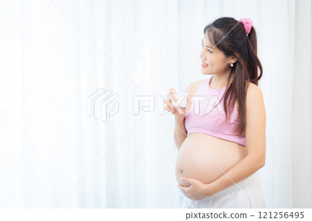 Asian pregnant woman relaxing at home.Asian pregnant woman is standing beside the window in living room and drinking a fresh milk in a glass. Asian pregnant woman portrait with copy space. Asian pregnant woman relaxing at home.Asian pregnant woman is standing beside the window in living room and drinking a fresh milk in a glass. Asian pregnant woman portrait with copy space. 121256495