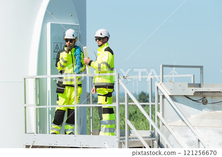Team of professional electrical engineers in fully safety suit are working and discussing together at the windmill electric generating turbine. Electricians working at the site of windmill turbine. Team of professional electrical engineers in fully safety suit are working and discussing together at the windmill electric generating turbine. Electricians working at the site of windmill turbine. 121256670
