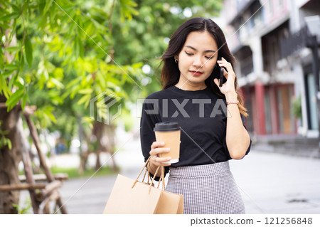 Smart and good looking Asian businesswoman is walking in the city while using a smartphone for text message and talking to colleague and other hand holding a coffee cup. Business woman portrait. Smart and good looking Asian businesswoman is walking in the city while using a smartphone for text message and talking to colleague and other hand holding a coffee cup. Business woman portrait. 121256848