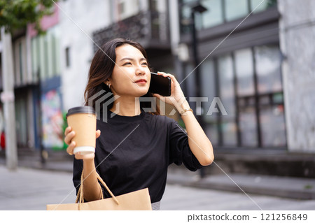 Smart and good looking Asian businesswoman is walking in the city while using a smartphone for text message and talking to colleague and other hand holding a coffee cup. Business woman portrait. Smart and good looking Asian businesswoman is walking in the city while using a smartphone for text message and talking to colleague and other hand holding a coffee cup. Business woman portrait. 121256849