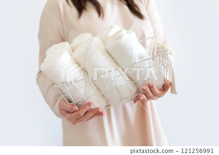 Female hands hold stylish white wire basket with white towels rolled into roll. Storage of towels. 121256991