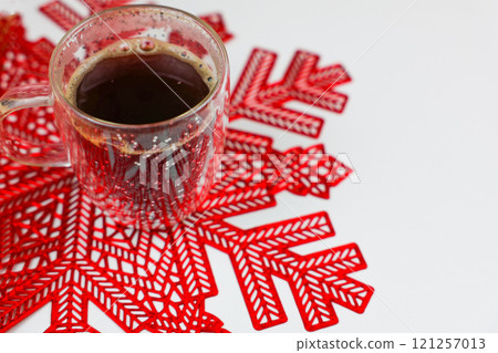 A transparent mug of coffee, in which foam and a bottle are visible, stands on a red napkin in the form of a snowflake on a white background in close-up with space for text. High quality photo A transparent mug of coffee, in which foam and a bottle are visible, stands on a red napkin in the form of a snowflake on a white background in close-up with space for text. High quality photo 121257013