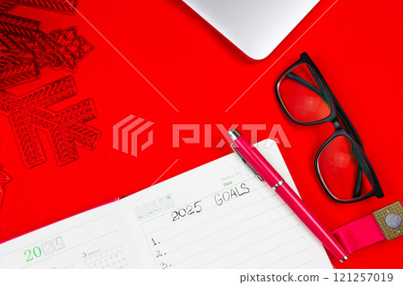 A pink notebook diary that says: Goals for 2025, inside it is a calendar for 2025 and there is a pink pen on a red background, a closed laptop lies on top, on the right there stands on a red New Year A pink notebook diary that says: Goals for 2025, inside it is a calendar for 2025 and there is a pink pen on a red background, a closed laptop lies on top, on the right there stands on a red New Year 121257019