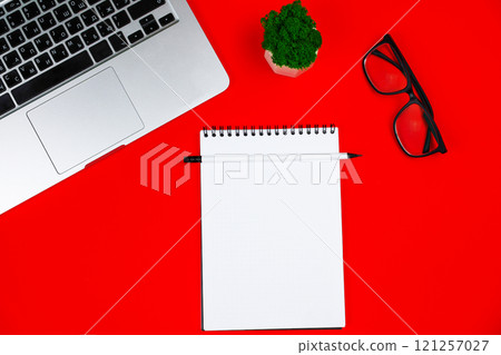 A white notebook with a black spring and a white pencil are lying on a red background, next to it are a gray laptop with a black keyboard, a green plant in a white pot: black-framed glasses and a red 121257027