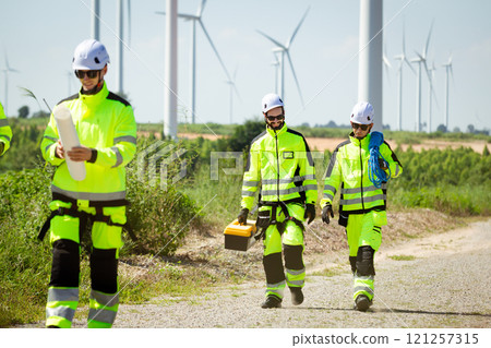 Team of professional electrical engineers in fully safety suit are working and discussing together at the windmill electric generating turbine. Electricians working at the site of windmill turbine. 121257315