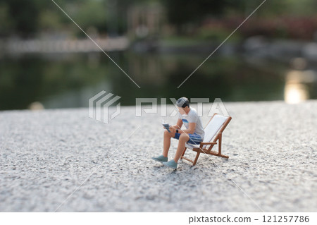 Miniature Person Relaxing on Deck Chair by Tranquil Lake Miniature Person Relaxing on Deck Chair by Tranquil Lake 121257786