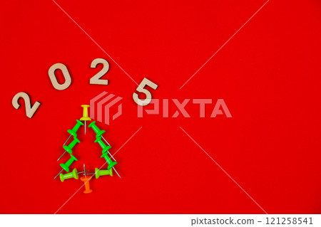 A creative Christmas tree consisting of green stationery pushpins stands on a red background, above it are wooden numbers 2025 with space for text. Office and holiday concept. High quality photo A creative Christmas tree consisting of green stationery pushpins stands on a red background, above it are wooden numbers 2025 with space for text. Office and holiday concept. High quality photo 121258541