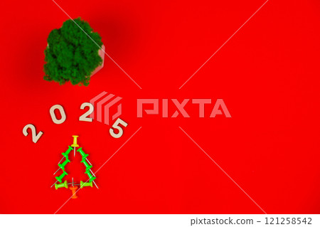 A creative Christmas tree consisting of green stationery pushpin on a bright red background, above it are wooden numbers 2025 and a green plant in a white pot with space for text. The concept of a A creative Christmas tree consisting of green stationery pushpin on a bright red background, above it are wooden numbers 2025 and a green plant in a white pot with space for text. The concept of a 121258542