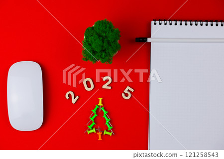 On a bright red background lies a creative Christmas tree made of office nails, above it are wooden numbers 2025, around it lie a white computer mouse, a green plant in a white pot, a white notebook On a bright red background lies a creative Christmas tree made of office nails, above it are wooden numbers 2025, around it lie a white computer mouse, a green plant in a white pot, a white notebook 121258543